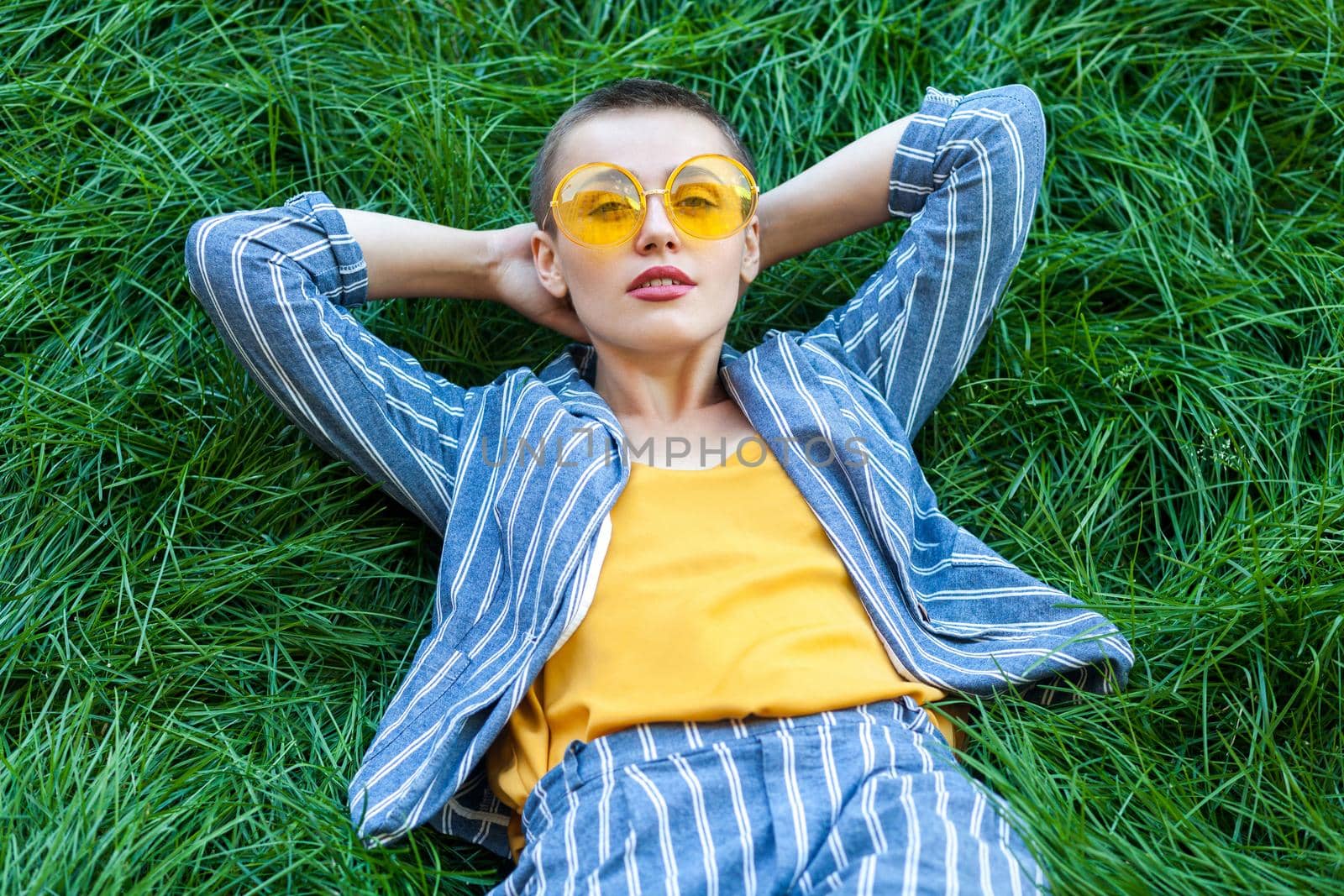 Portrait of beautiful young woman with short hair in casual blue striped suit, yellow shirt, glasses lying down on green grass, resting, holding hand behind head and looking at camera. summertime shot