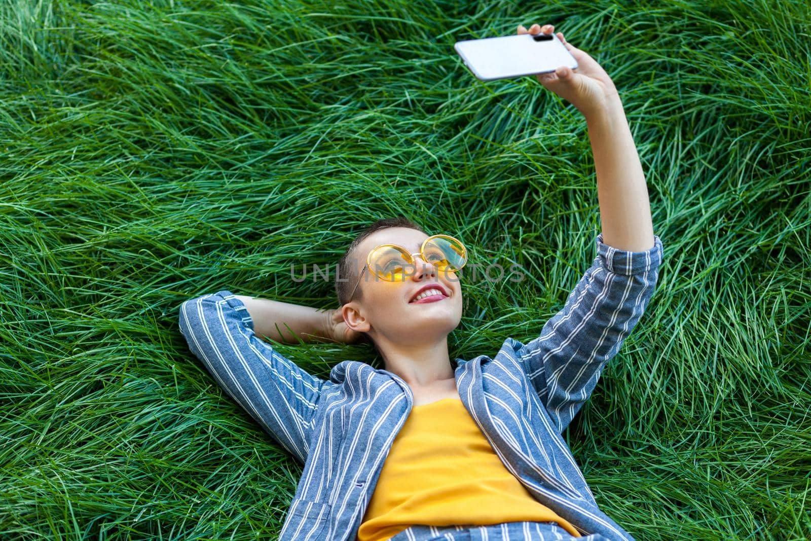 Portrait of smiley young short hair woman in casual blue striped suit, yellow shirt, glasses lying down on green grass holding her mobile phone, doing selfie or online video. outdoor summertime shot.