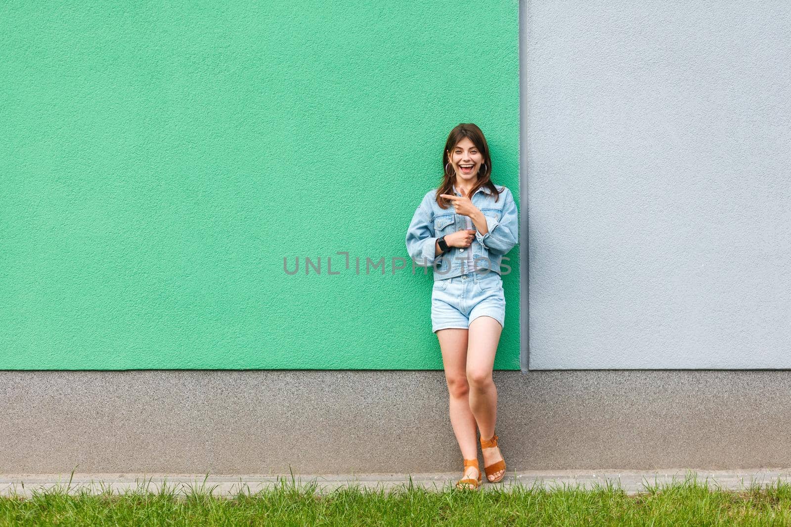 Full length portrait of happy excited beautiful woman in casual jeans denim style in summertime standing near green and light blue wall and pointing at green copyspace. two different choice concept.