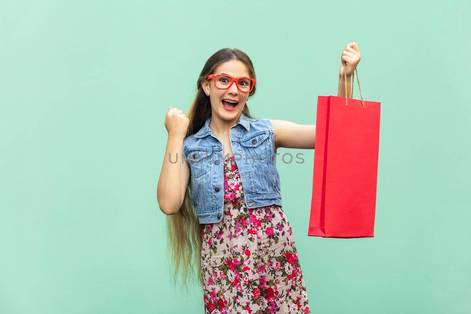 The beautiful long haired girl in casual clothing with shopping bags, looking at camera, celebrates the victory. Blonde model. Sales, shop, retail, consumer concept. Isolated studio shot on light blue background