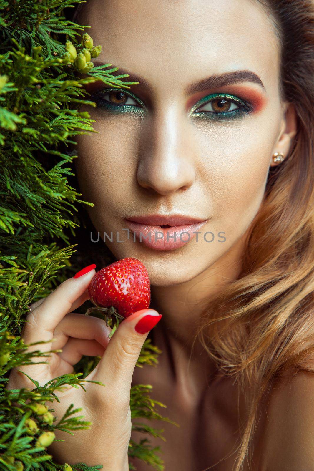 Handsome model with perfect green makeup looking at camera and holding red berry. Outdoor spring or summer photo