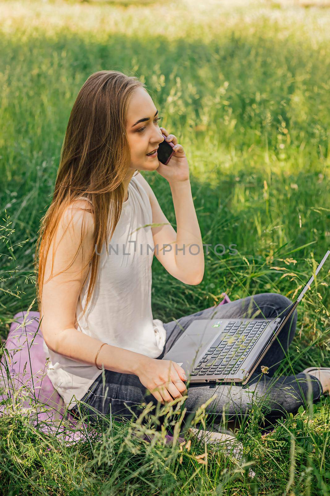 girl sits on the grass and works at a laptop. talking on the phone. freelance. selfeducation. the concept of remote learning and outdoor work. by Anyatachka