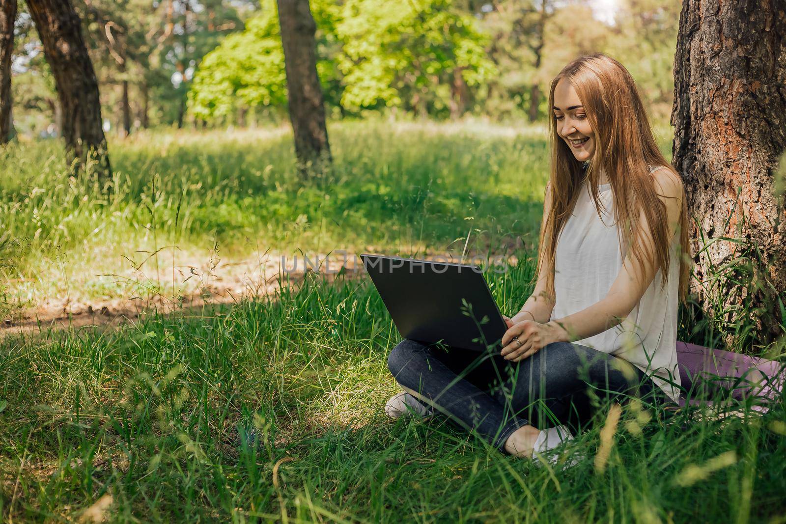On the banner, a young girl works with a laptop in the fresh air in the park, sitting on the lawn. The concept of remote work. Work as a freelancer. The girl takes courses on a laptop and smiles. by Anyatachka