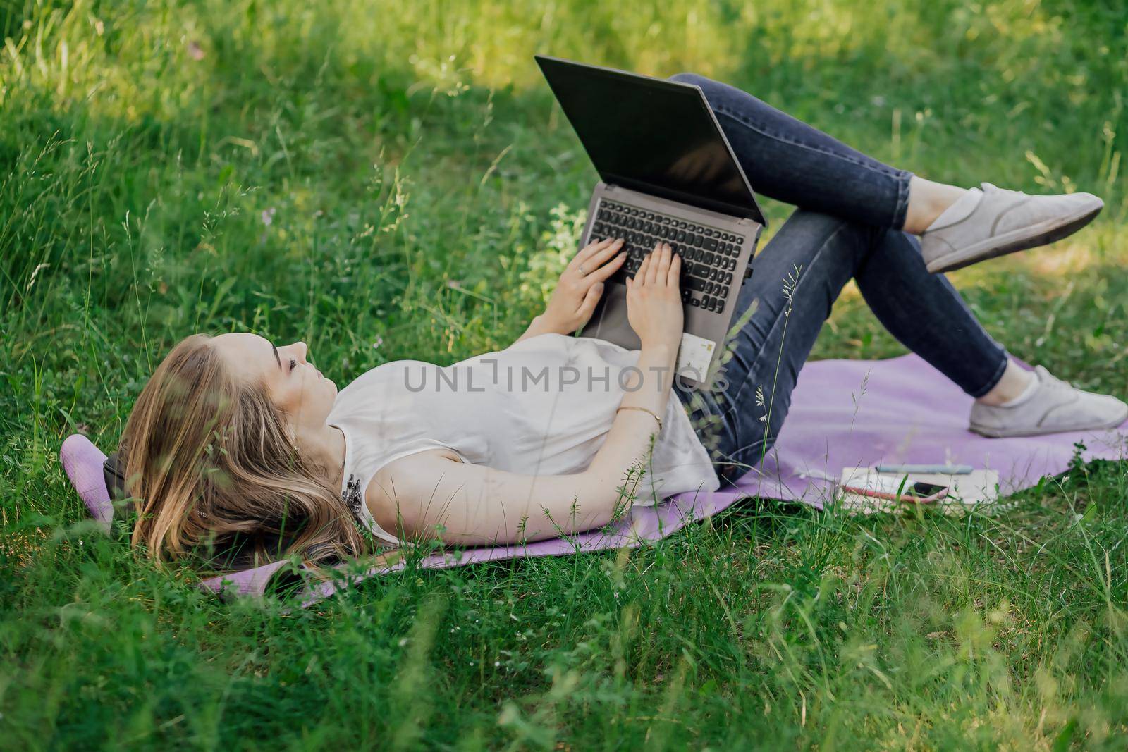 On the banner, a young girl works with a laptop in the fresh air in the park, sitting on the lawn. The concept of remote work. Work as a freelancer. The girl takes courses on a laptop and smiles. by Anyatachka