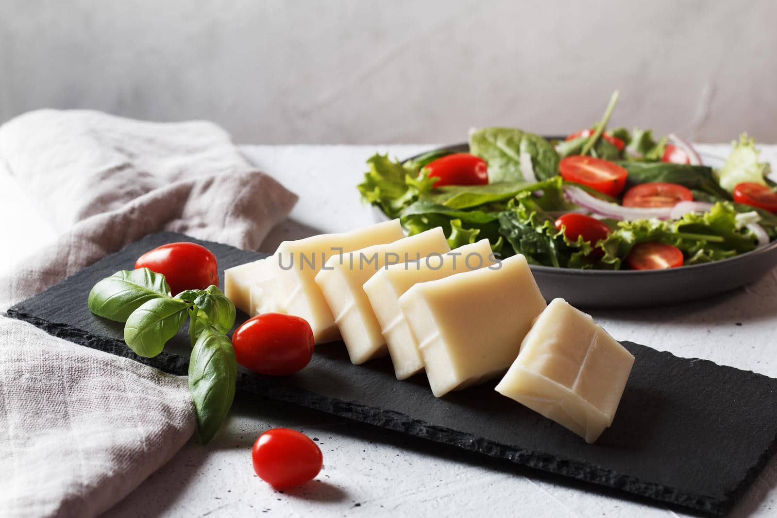 A piece of fresh sliced halloumi on a slate board with cherry tomatoes and spinach leaves. Preparing to cook cheese on the grill.