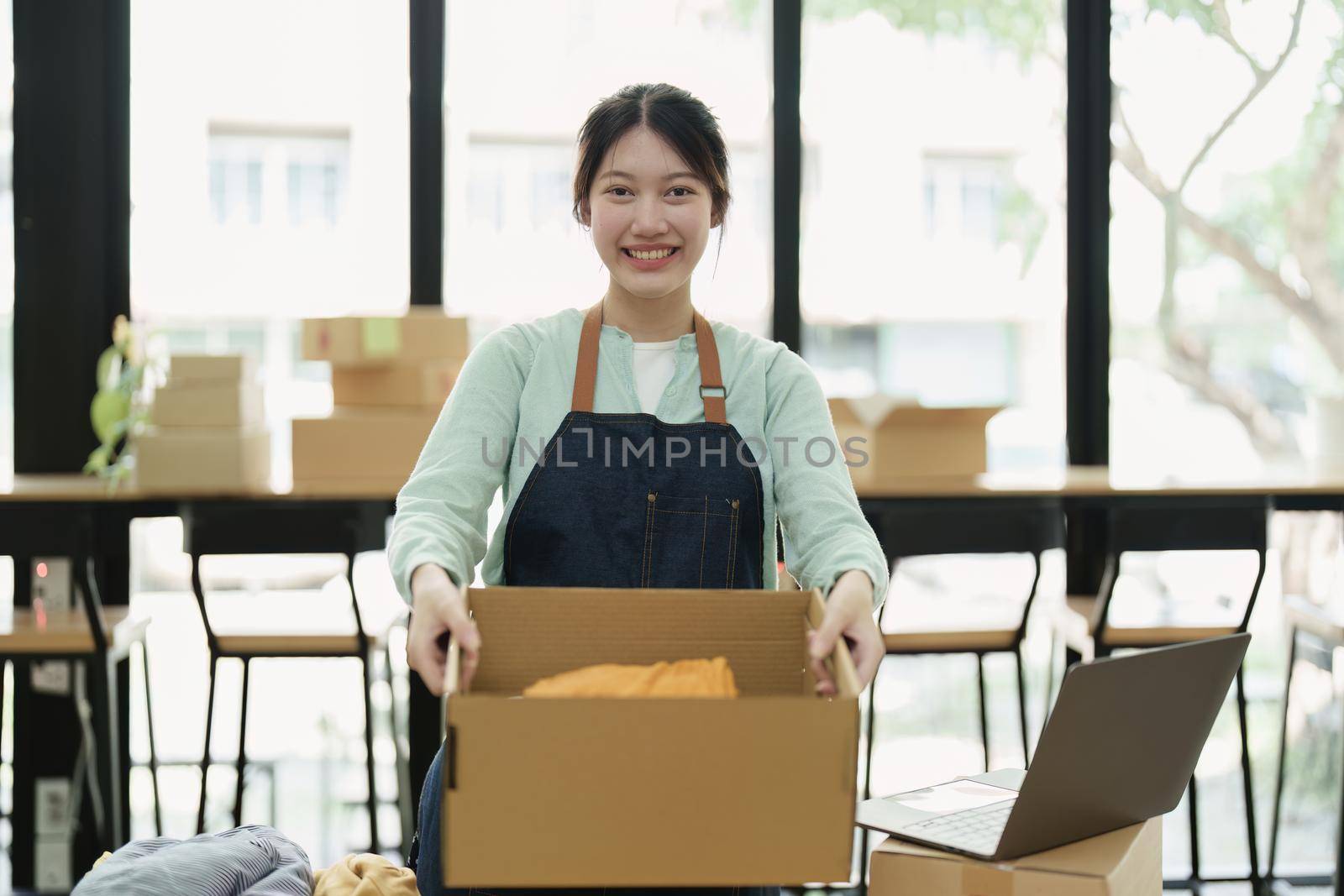 Portrait of a small startup Asian female entrepreneur SME owner picking up a yellow shirt before packing it in an inner box with a customer. Online Business Ideas and Freelance.