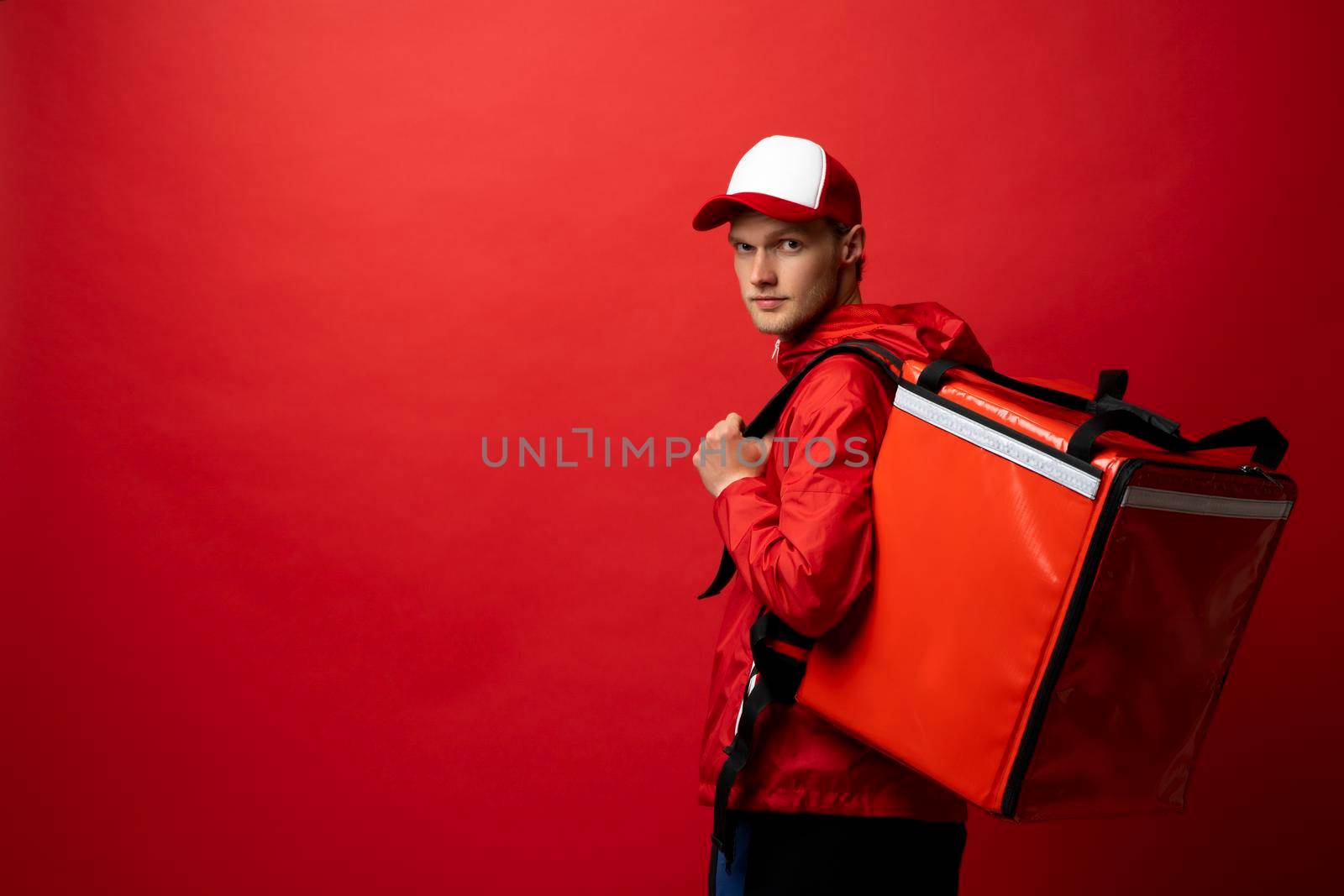 Delivery guy employee man in red cap T-shirt uniform work as dealer courier hold thermal food bag backpack isolated on red background