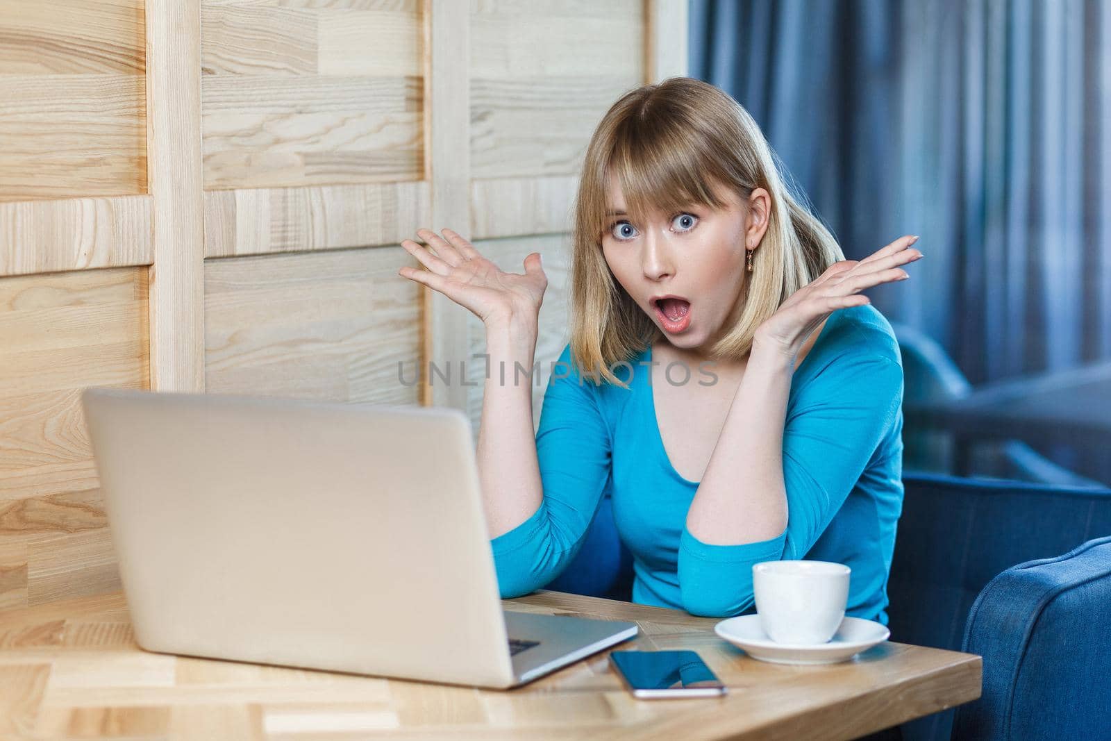 Wow! Portrait of emotional shocked young businesswoman in blue blouse are sitting in cafe, reading news and remotely working with suprised big eyes, open mouth and raised arms looking at camera.Indoor