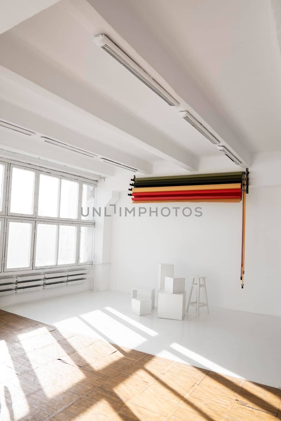 Sunny daylight photo studio interior with set of professional equipment as a rolls of color background and white cubes on a white floor