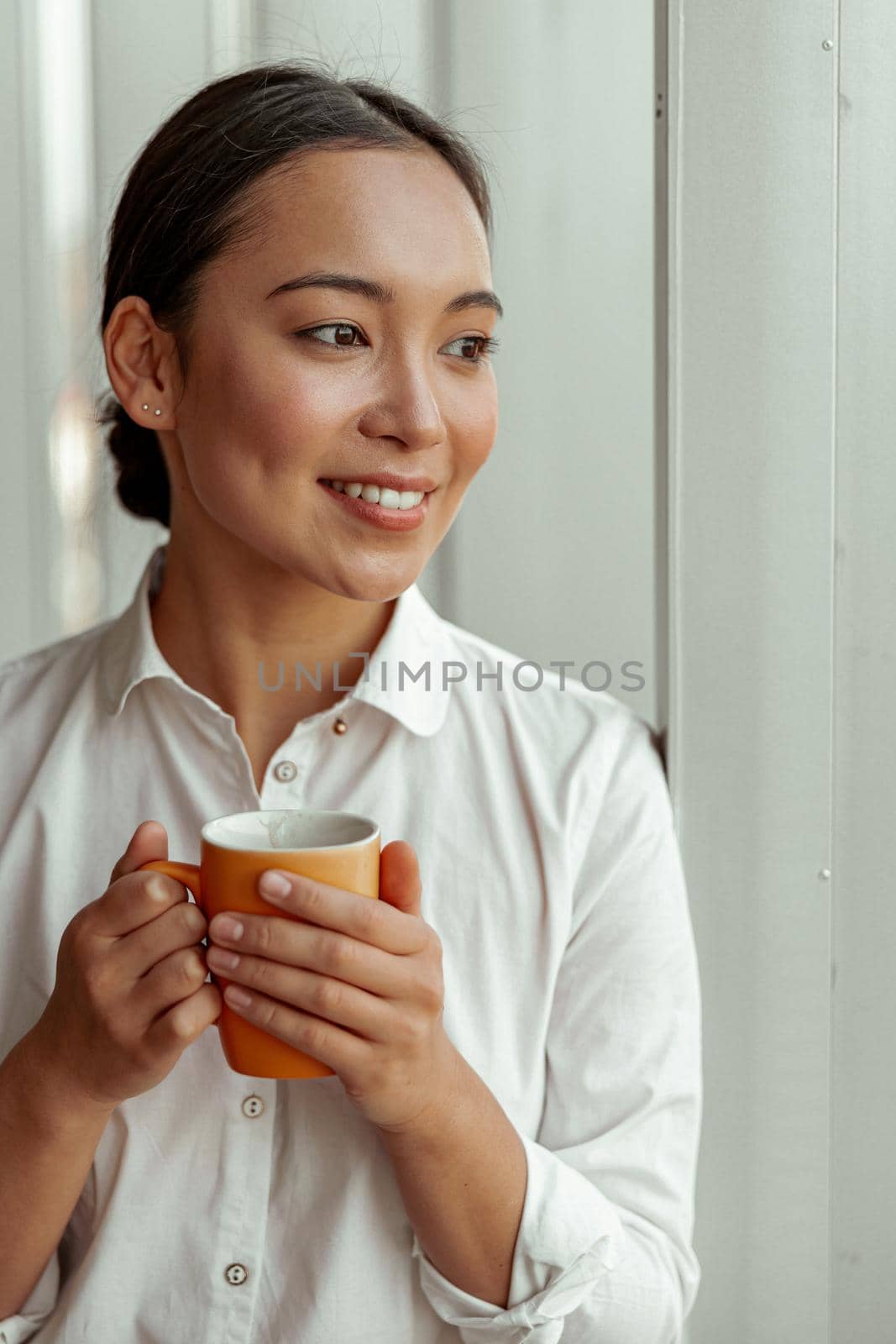 Portrait of charming asian business woman drinking coffee standing near windows at office by Yaroslav_astakhov