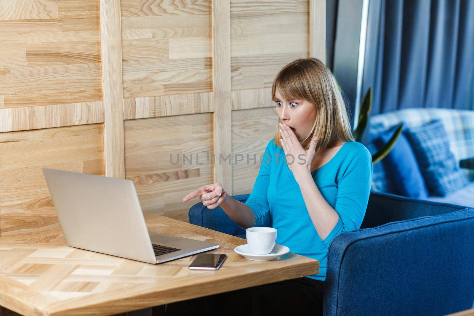 No way! Side view portrait of unbelievable young girl with blonde hair in blue t-shirt are sitting in cafe, working and making video call on laptop, pointing finger to the companion with shocked face