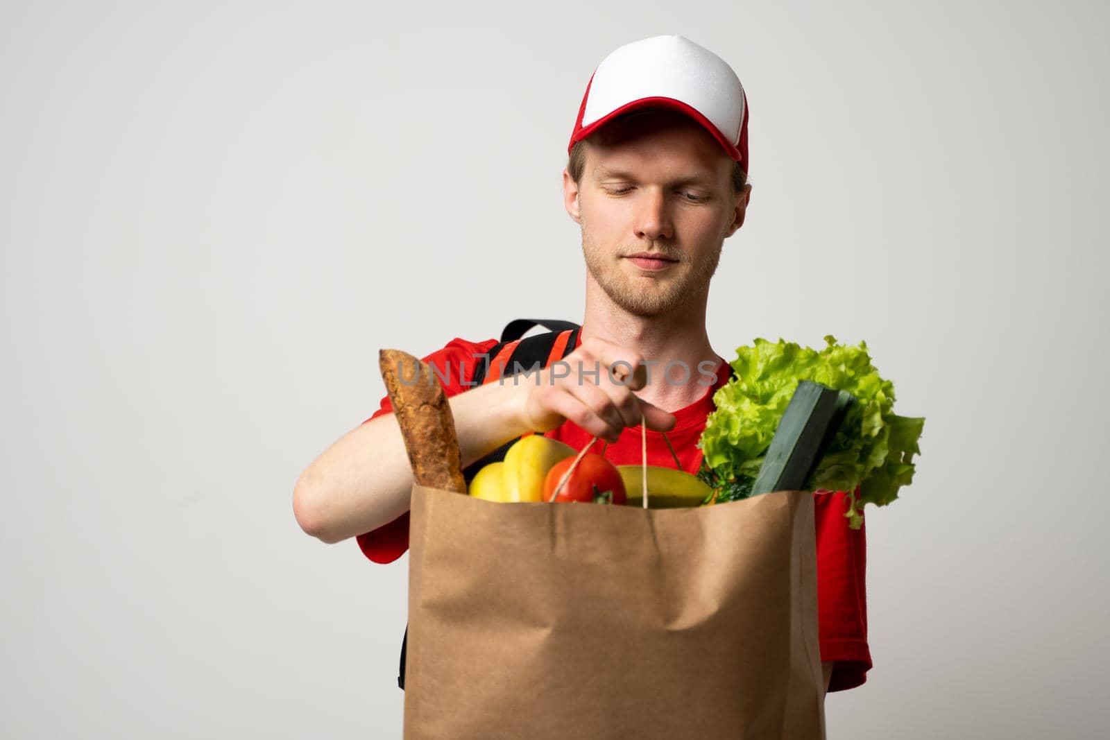 Grocery delivery courier man in a red cap and t-shirt with a thermal bag on a shoulders holds paper bag with food on white studio background. Food delivery service, online shopping