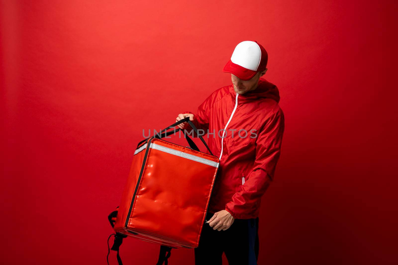 Professional delivery employee man 20s in red cap T-shirt uniform workwear work as dealer courier with red thermal food bag backpack isolated on red background. Service concept. by vovsht