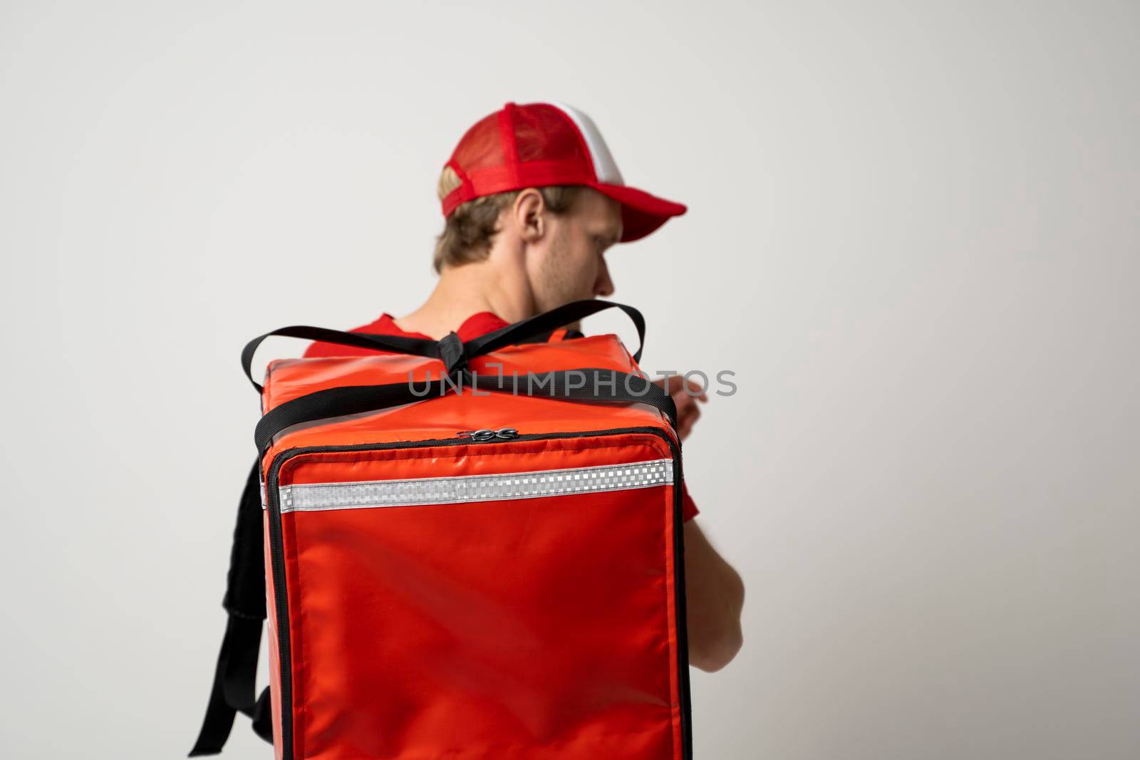 Delivery guy employee man in red cap T-shirt uniform work as dealer courier and hold red thermal food bag backpack. by vovsht