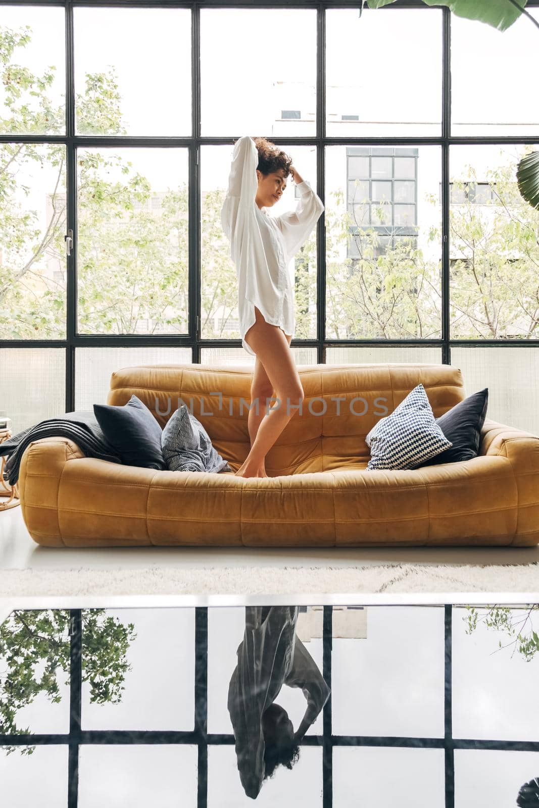 Pensive young african american latina woman standing on couch in loft apartment. Vertical image. Lifestyle concept.