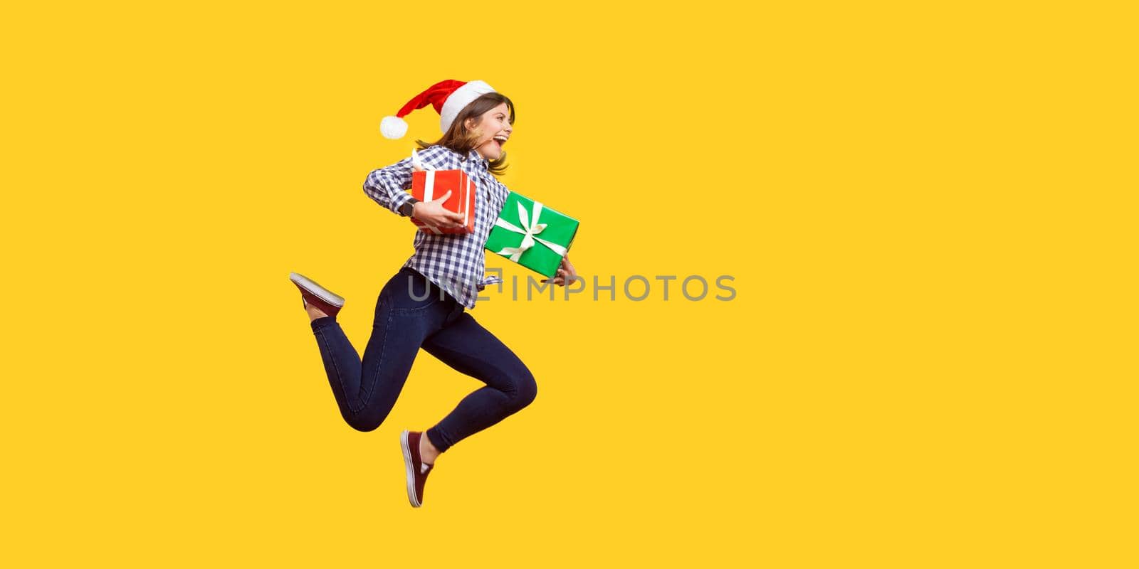 Side view portrait of extremely joyous brunette woman in santa hat jumping or running in air with wrapped xmas gift boxes, celebrating discounts. indoor studio shot isolated on yellow background