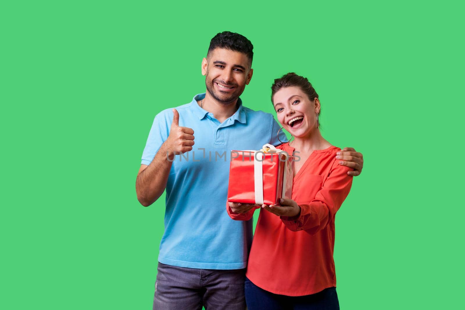 Like this present. Portrait of cheerful young couple in casual wear standing together, woman showing gift box to camera, man gesturing thumbs up. isolated on green background, indoor studio shot