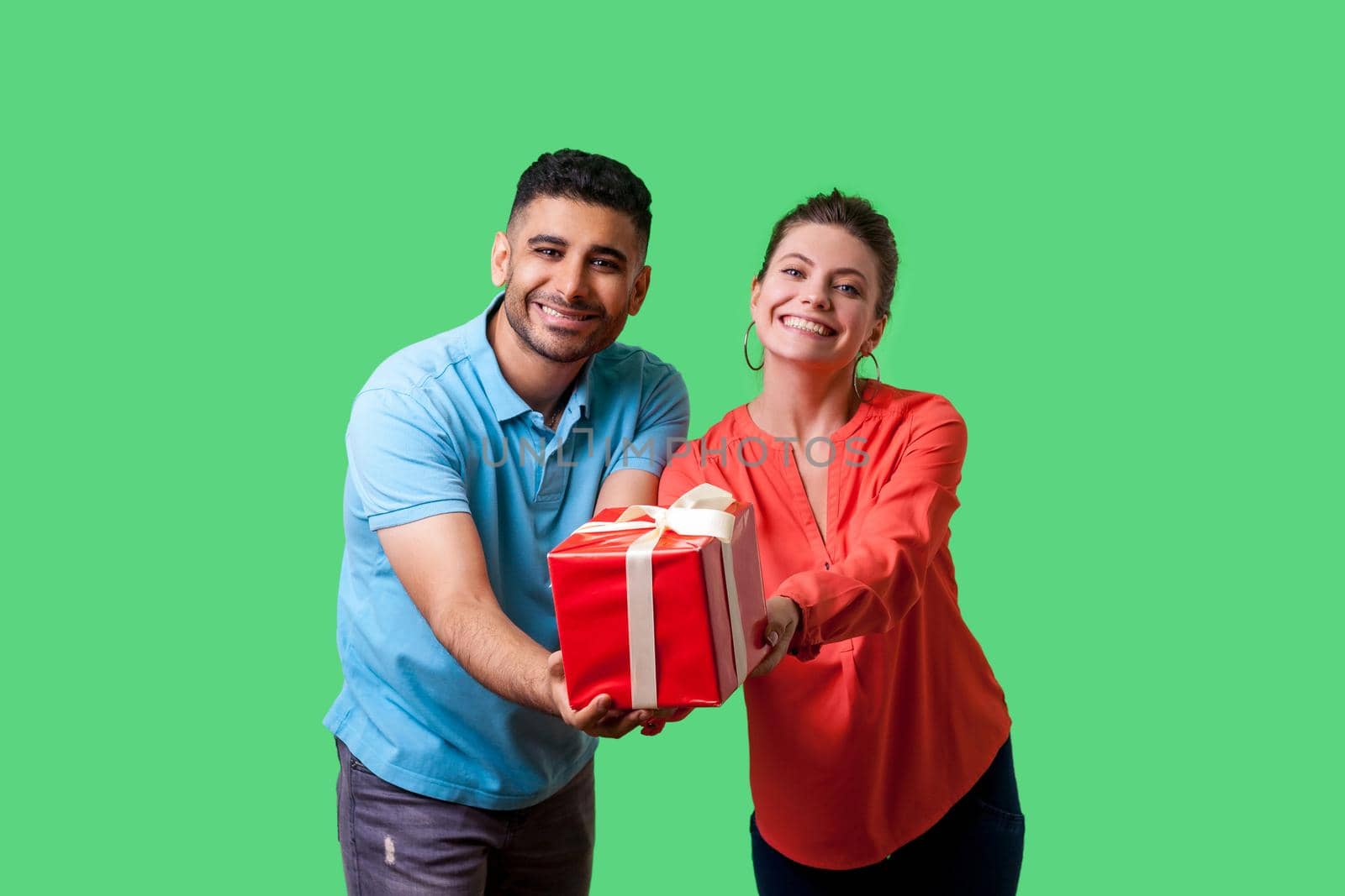 Please take your present. Young generous couple giving gift box to camera, looking with toothy smile, sharing present on holidays, charity concept. isolated on green background, indoor studio shot