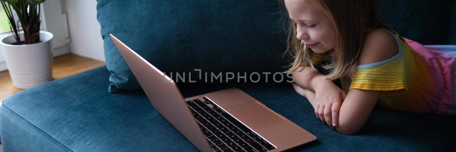 A little girl is lying on the couch with a laptop by kuprevich
