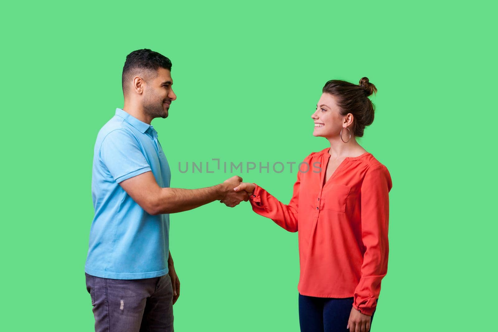 Portrait of positive friendly man and woman in casual wear standing, smiling at each other and shaking hands, couple first meeting or acquaintance. isolated on green background, indoor studio shot