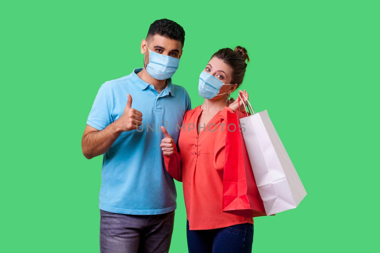 Portrait of satisfied young couple with surgical medical masks showing thumbs up gesture together, cute woman holding bags and smiling. isolated on green background, indoor studio shot