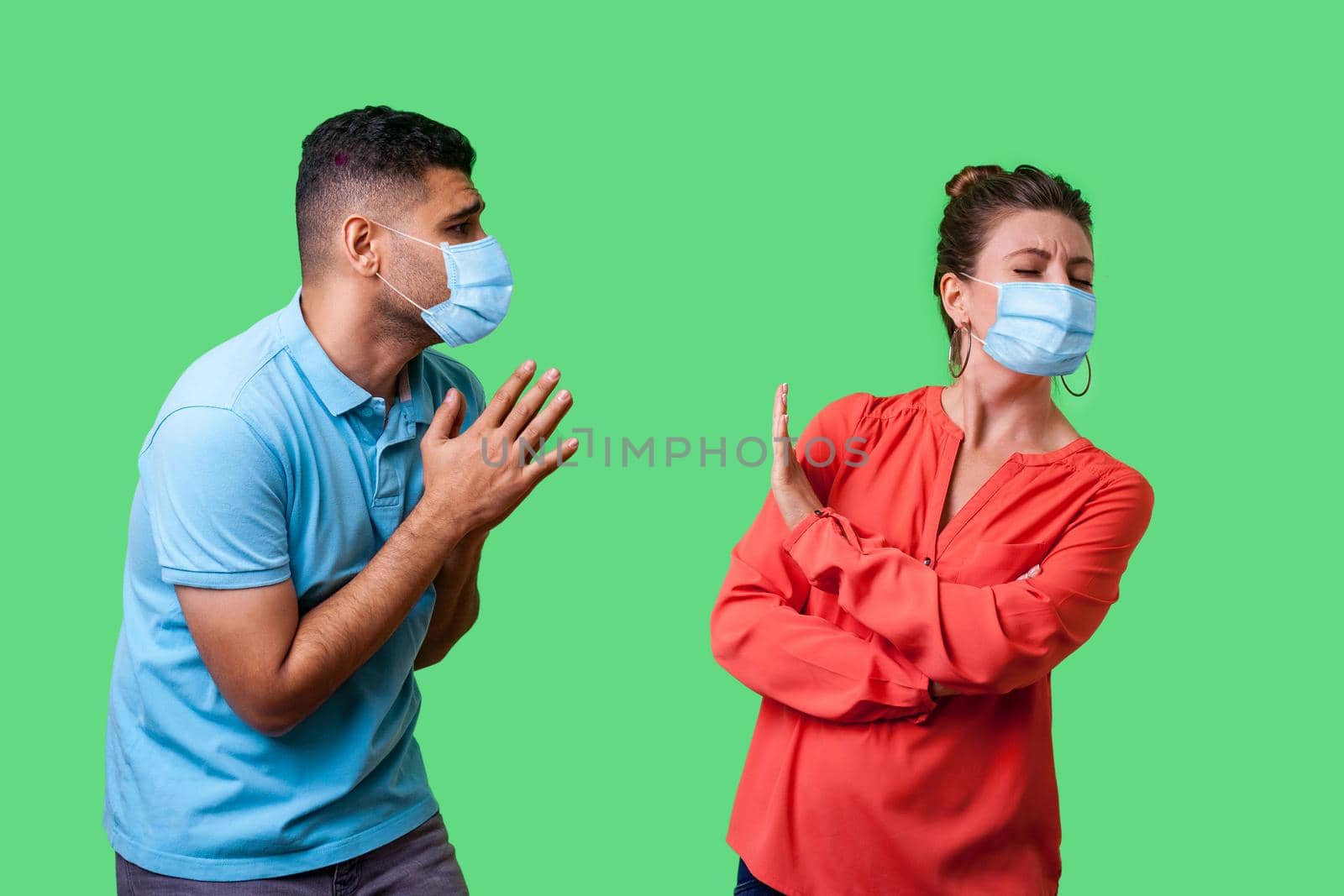 Portrait of couple with surgical medical mask standing together, man holding arms in prayer and pleading forgiveness, looking with hope at naughty woman, isolated on green background, indoor shot