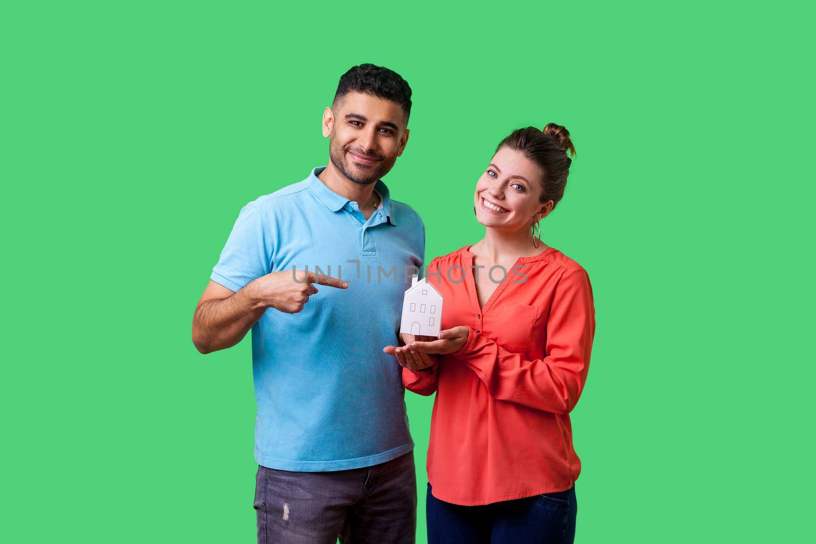 Portrait of happy couple in casual clothes looking at camera with toothy smile, man pointing at paper house in woman hands, dreaming of own home. isolated on green background, indoor studio shot