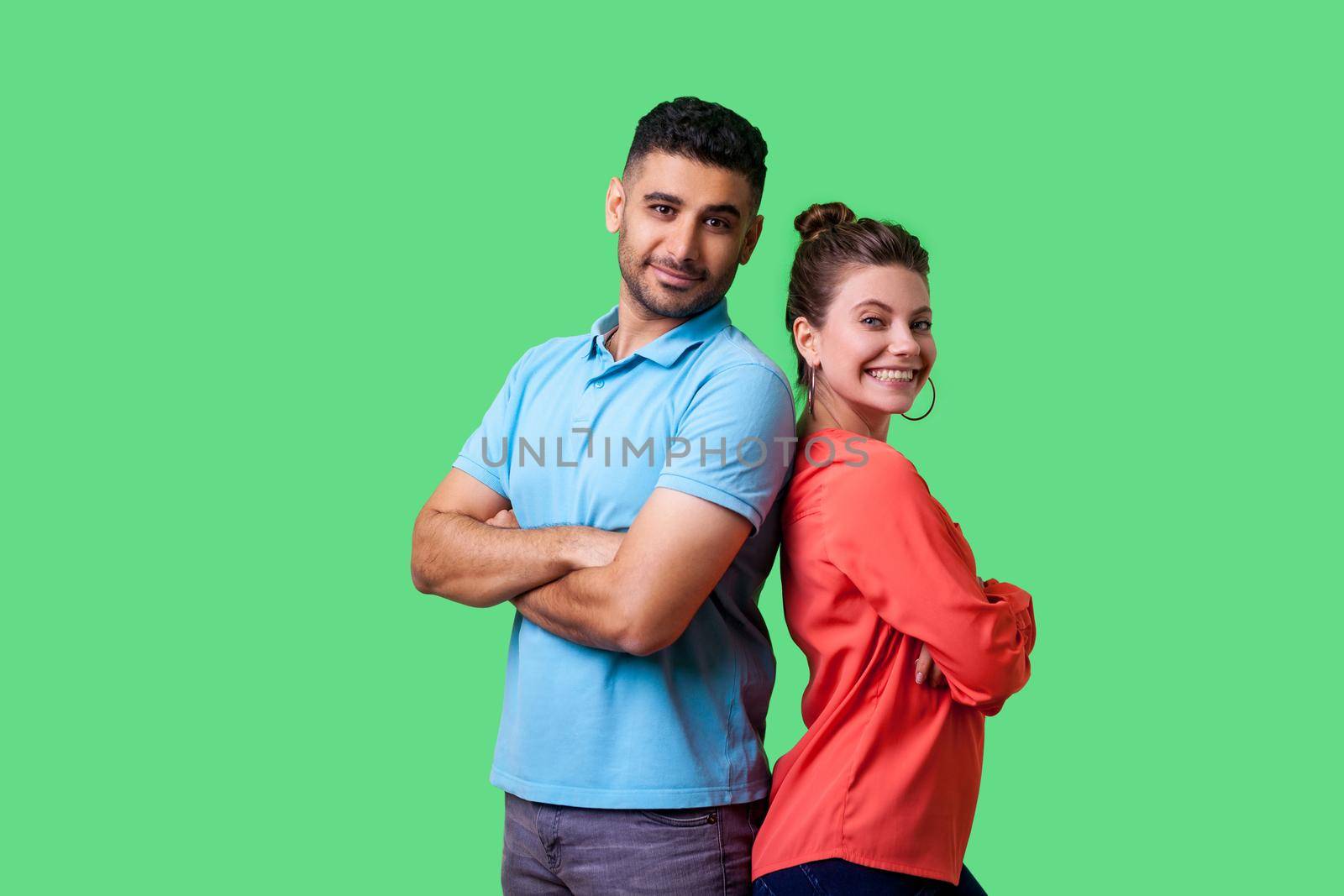 Reliable friendship. Cheerful young couple of friends in casual wear standing back to back with crossed hands and looking at camera with toothy smile. isolated on green background, indoor studio shot