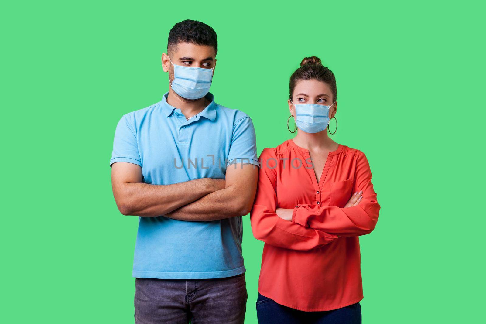 Portrait of upset couple with surgical medical mask standing together with crossed hands, looking sideways at each other with resentful glance, suspicion. isolated on green background, indoor studio