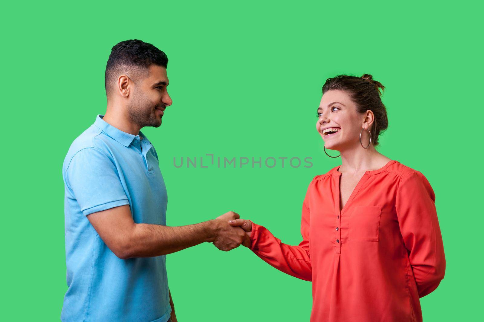 Nice to meet you. Portrait of happy young couple in casual wear standing, smiling at each other and shaking hands, friendly meeting or acquaintance. isolated on green background, indoor studio shot