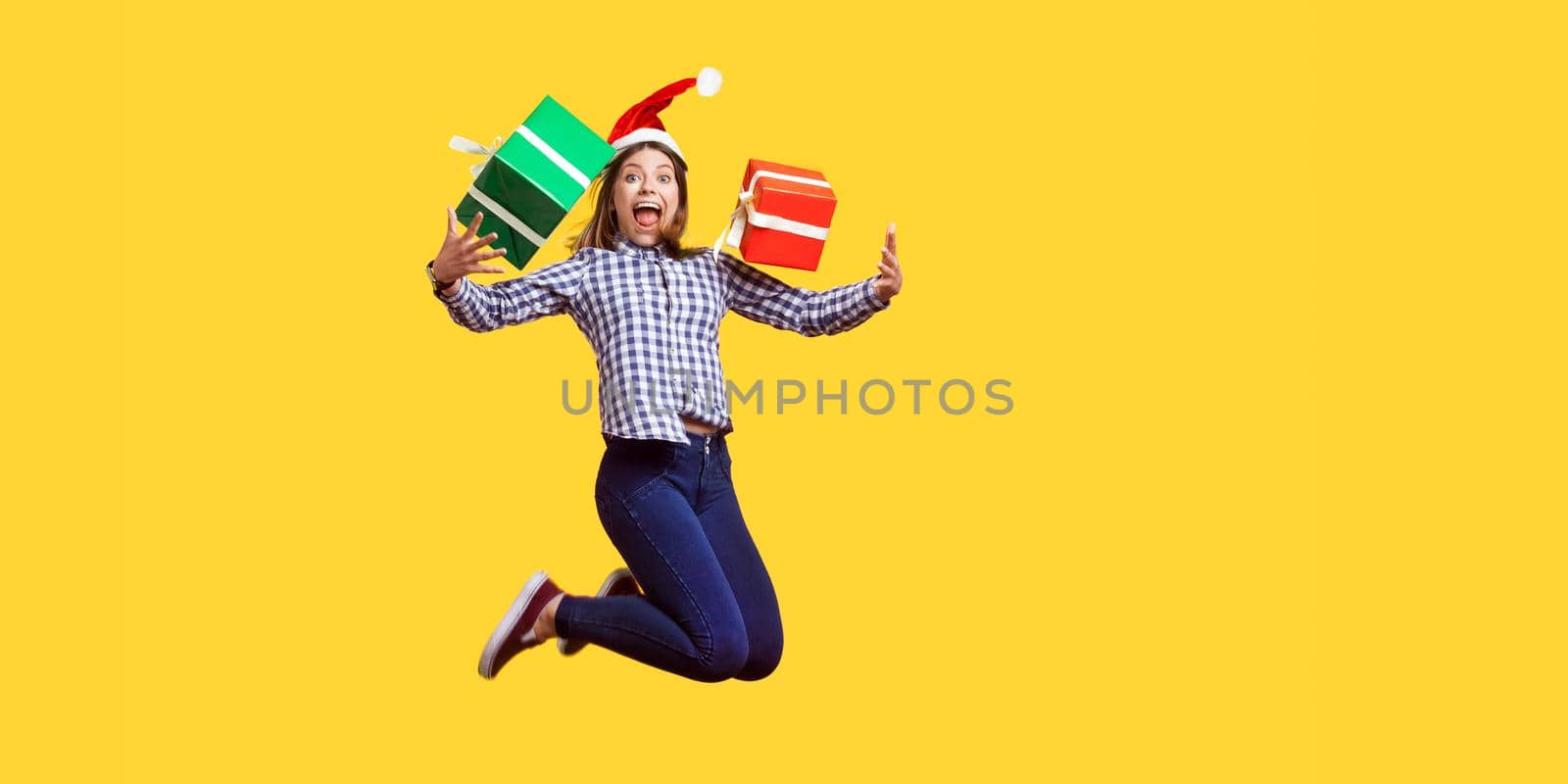 Portrait of extremely happy brunette woman in santa hat and checkered shirt jumping for joy, flying with wrapped xmas gift boxes, celebrating sales. indoor studio shot isolated on yellow background