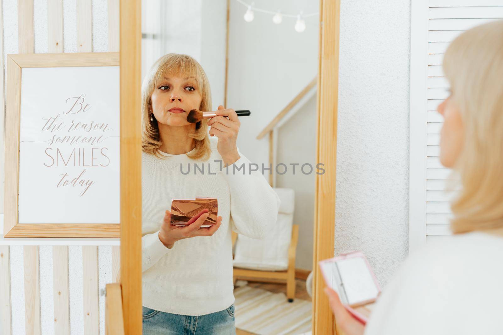 Beautiful smiling mature senior caucasian woman with blonde hair standing near mirror with make up palette and brush, doing makeup and looking at reflection in living room at home by Ostanina