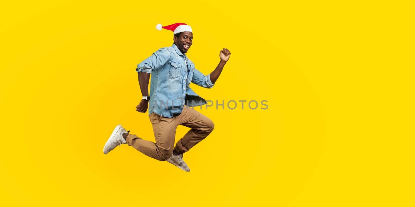 Full length portrait of happy joyous man with new year santa hat jumping or flying, hurry running to his dream, looking at camera with toothy smile. indoor studio shot isolated on yellow background