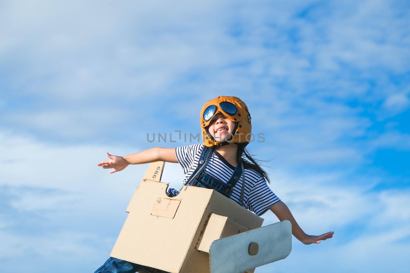 Cute dreamer little girl playing with cardboard planes against blue summer sky background. Happy kid running through the meadow on a sunny day with cardboard plane.  Childhood dream imagination concep by TEERASAK