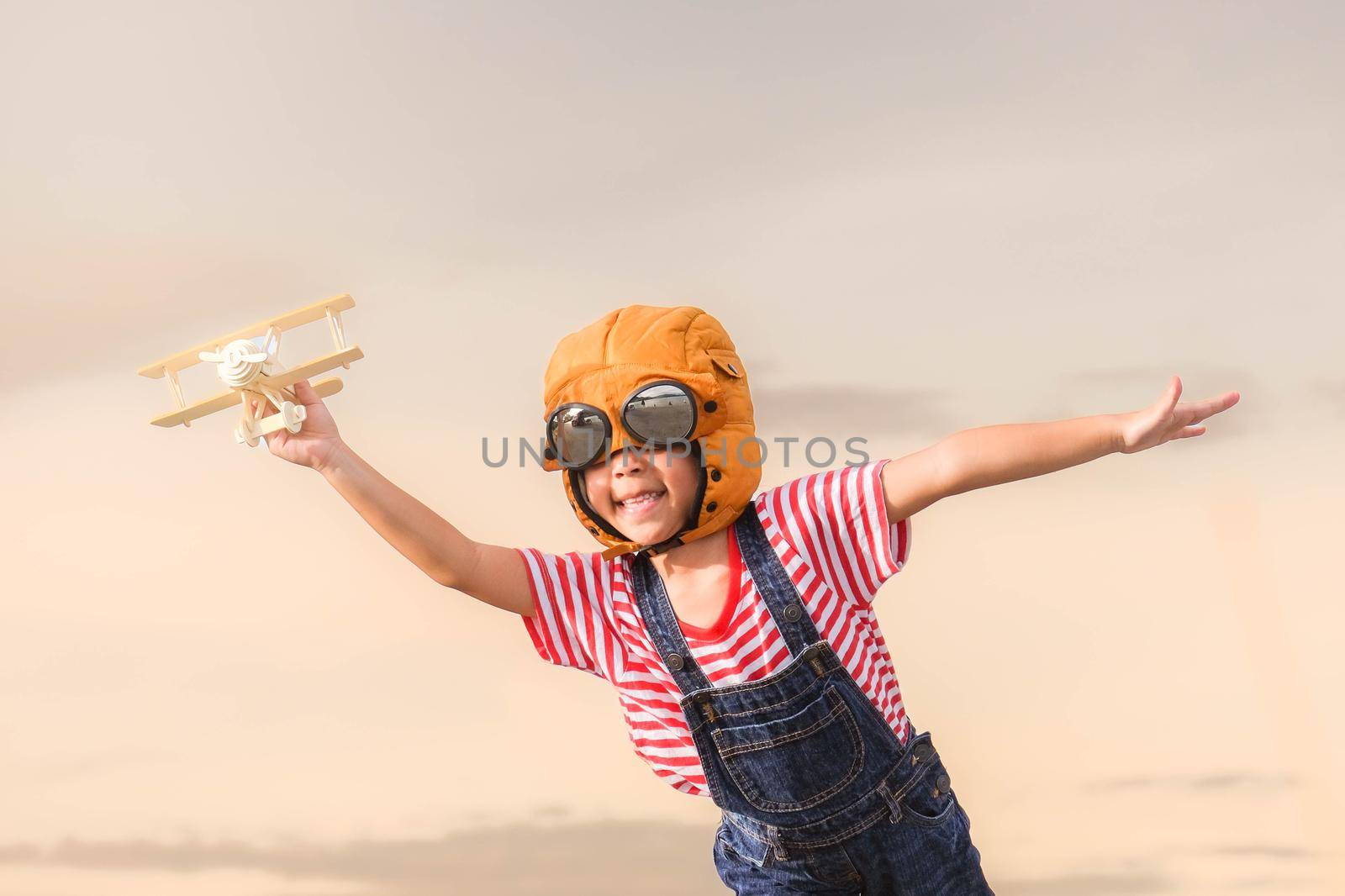 Cute little girl playing with toy plane against pastel sky background. Happy kid in pilot hat playing with toy plane in summer park. Childhood dream imagination concept.