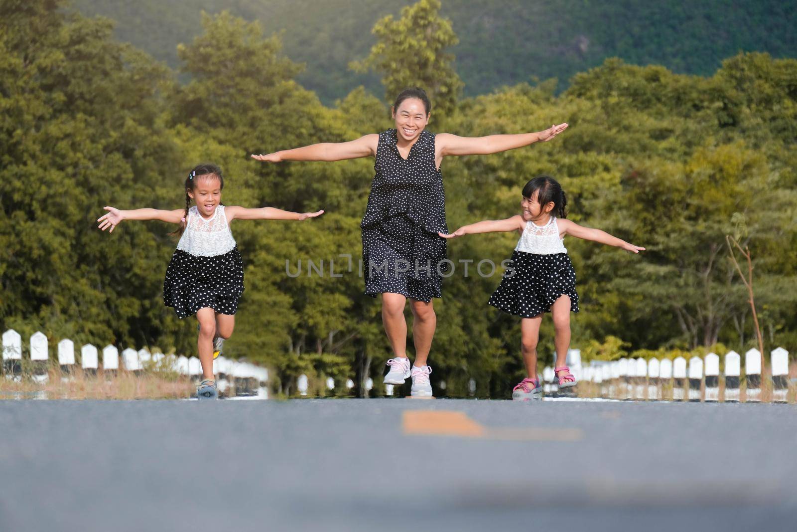 Two lovely daughters are running with their mother spreading their arms as if flying on the road in the park. Happy mother plays with the children in the park, spending time together on vacation.