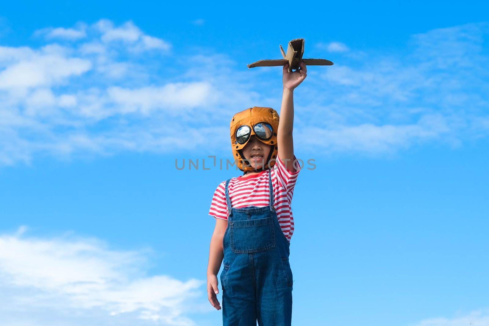Cute little girl running through the meadow on a sunny day with a toy plane in hand. Happy kid playing with cardboard plane against blue summer sky background. Childhood dream imagination concept. by TEERASAK