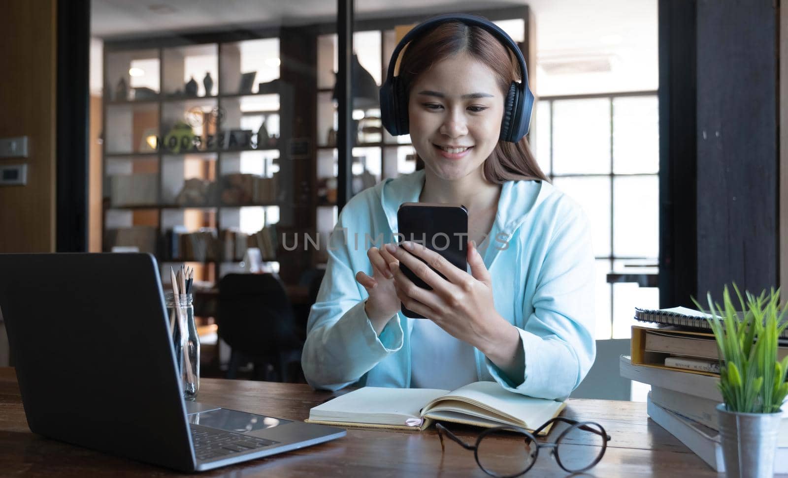 Beautiful millennial Asian female university student listening to music with her modern wireless headphones and using smartphone. by wichayada