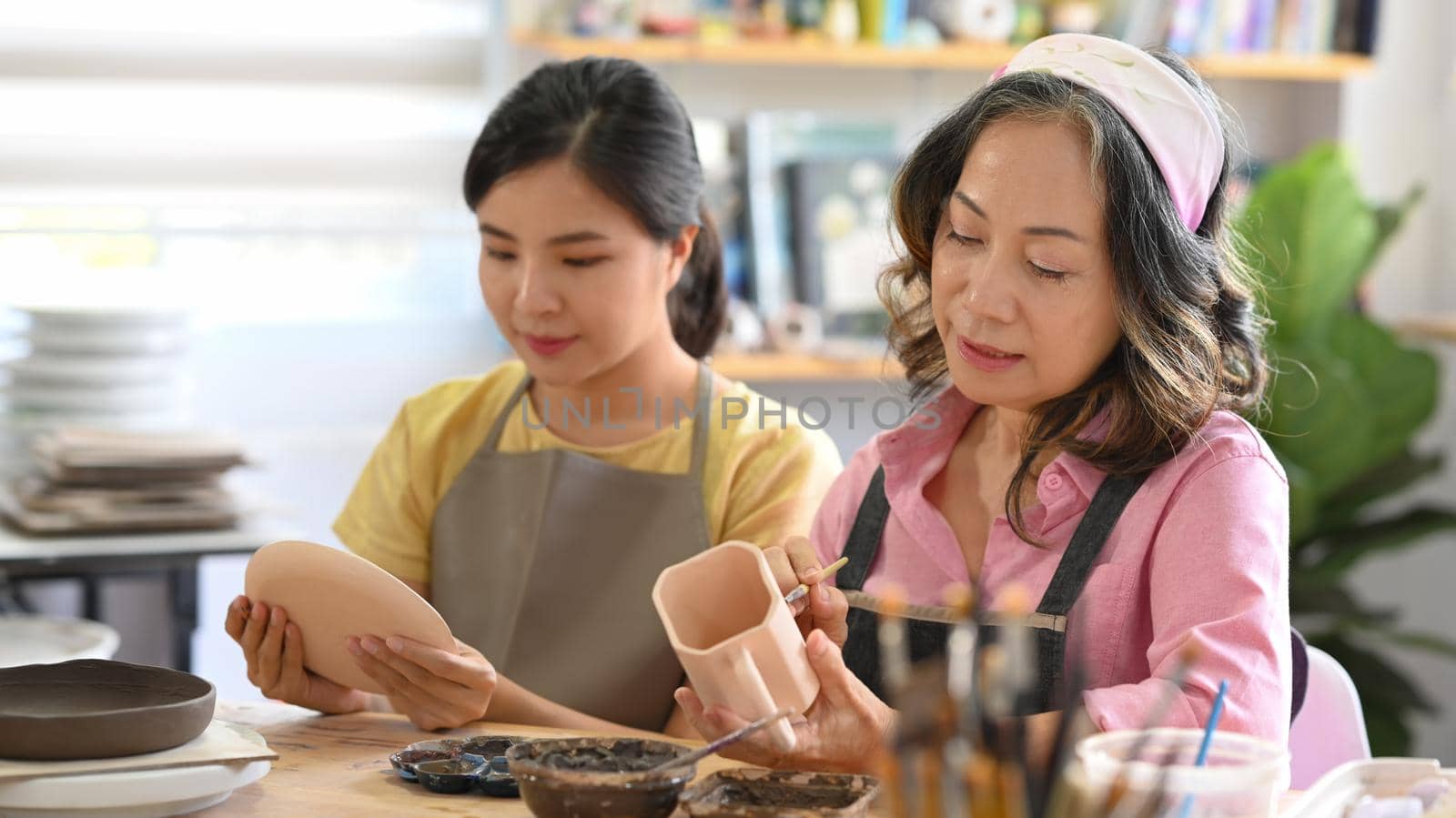 Attractive mature woman and young woman painting pottery bowl in workshop. Indoors lifestyle activity, handicraft, hobbies concept.