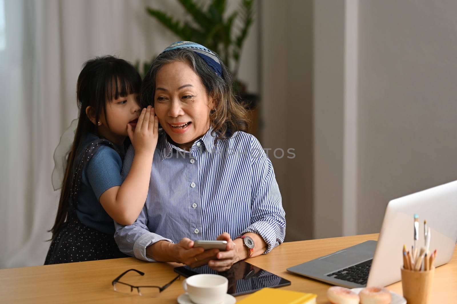 Adorable little girl whispering something to happy senior grandmother, spending leisure weekend at home together. Loving family relationship concept.