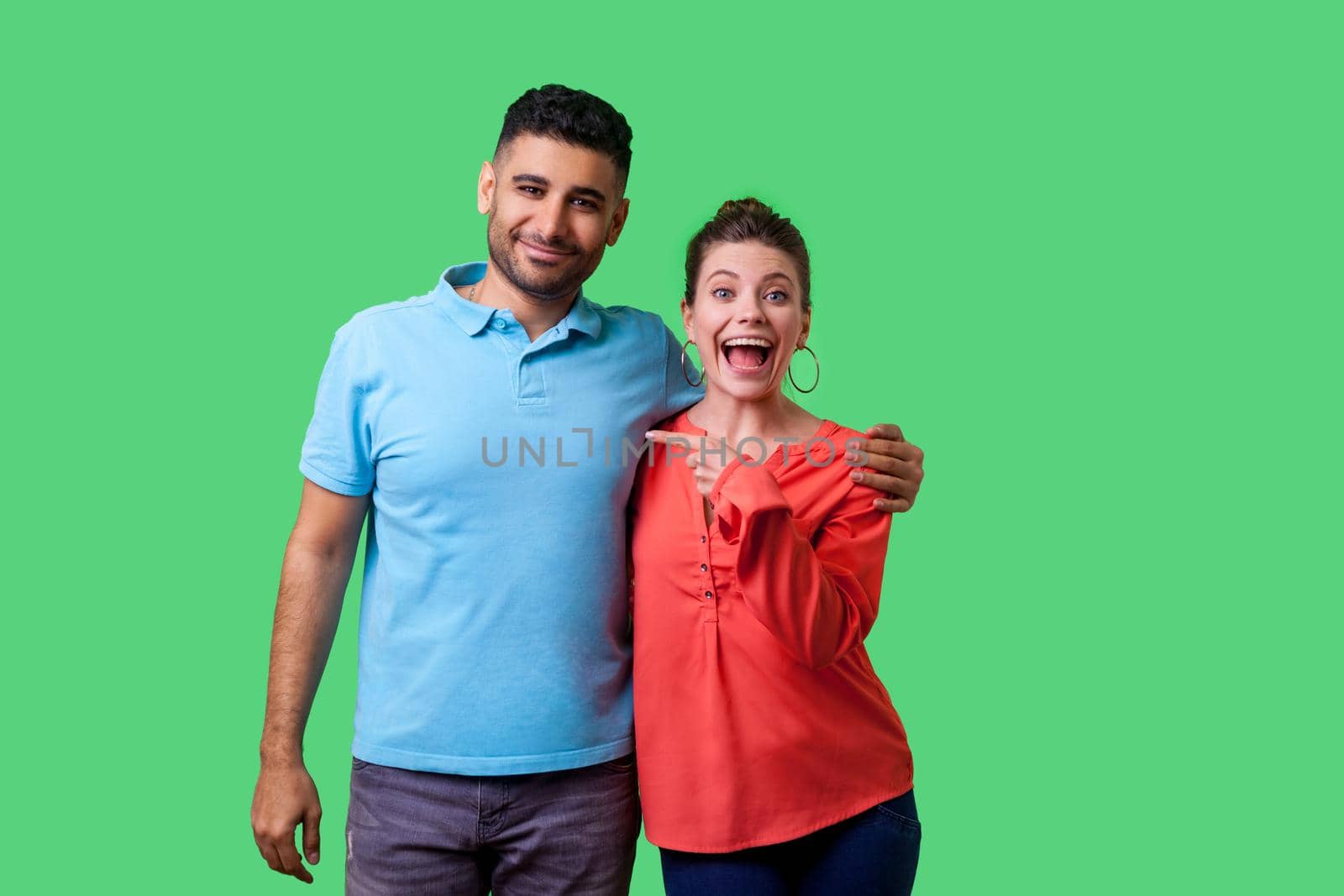That's him. Portrait of happy young couple in casual wear standing and hugging as friends, woman pointing finger at man, looking shocked at camera. isolated on green background, indoor studio shot