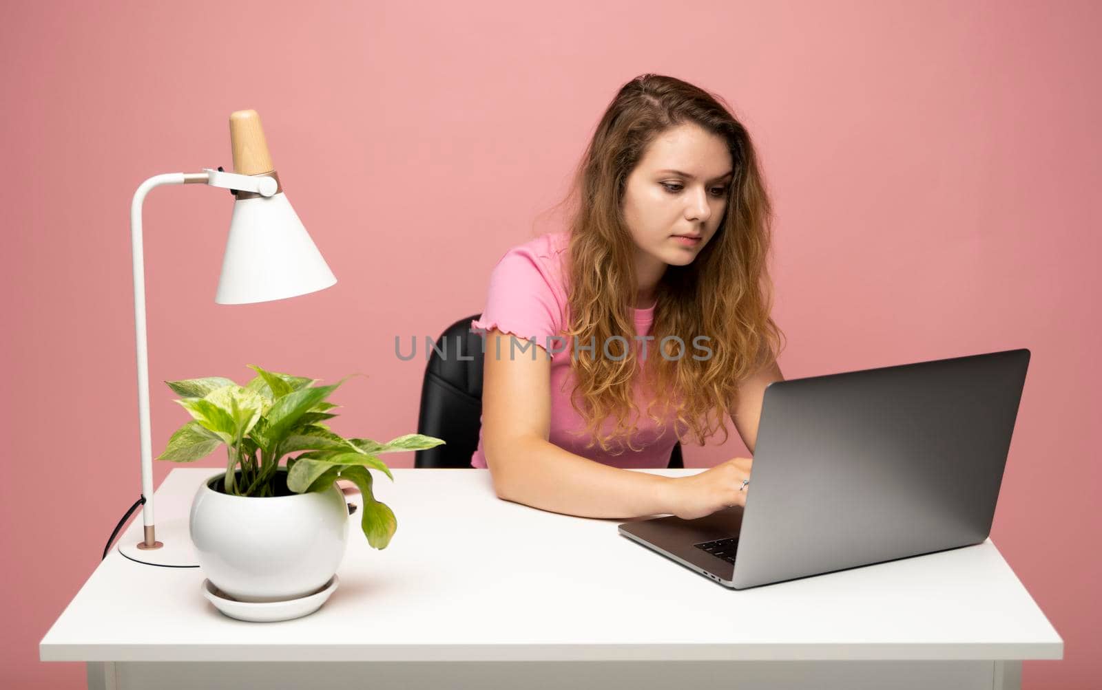 Young freelancer curly woman in a pink t-shirt working with a laptop computer. Working on a project. Freelance worker. by vovsht