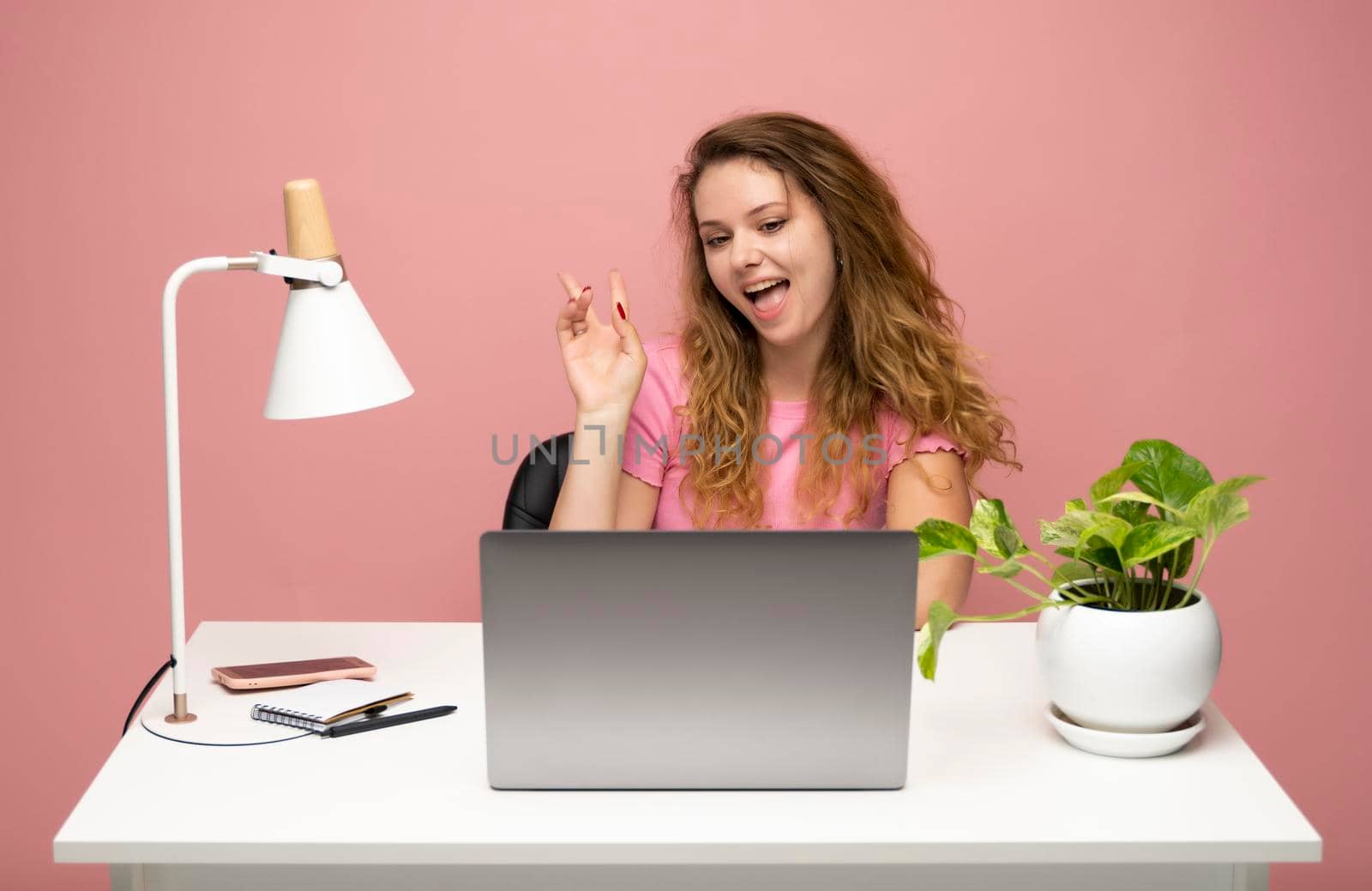 Happy young freelancer curly woman in a blue t-shirt working with a laptop computer and speaking with a client or friends with messengers. Working on a project. Freelance worker. by vovsht