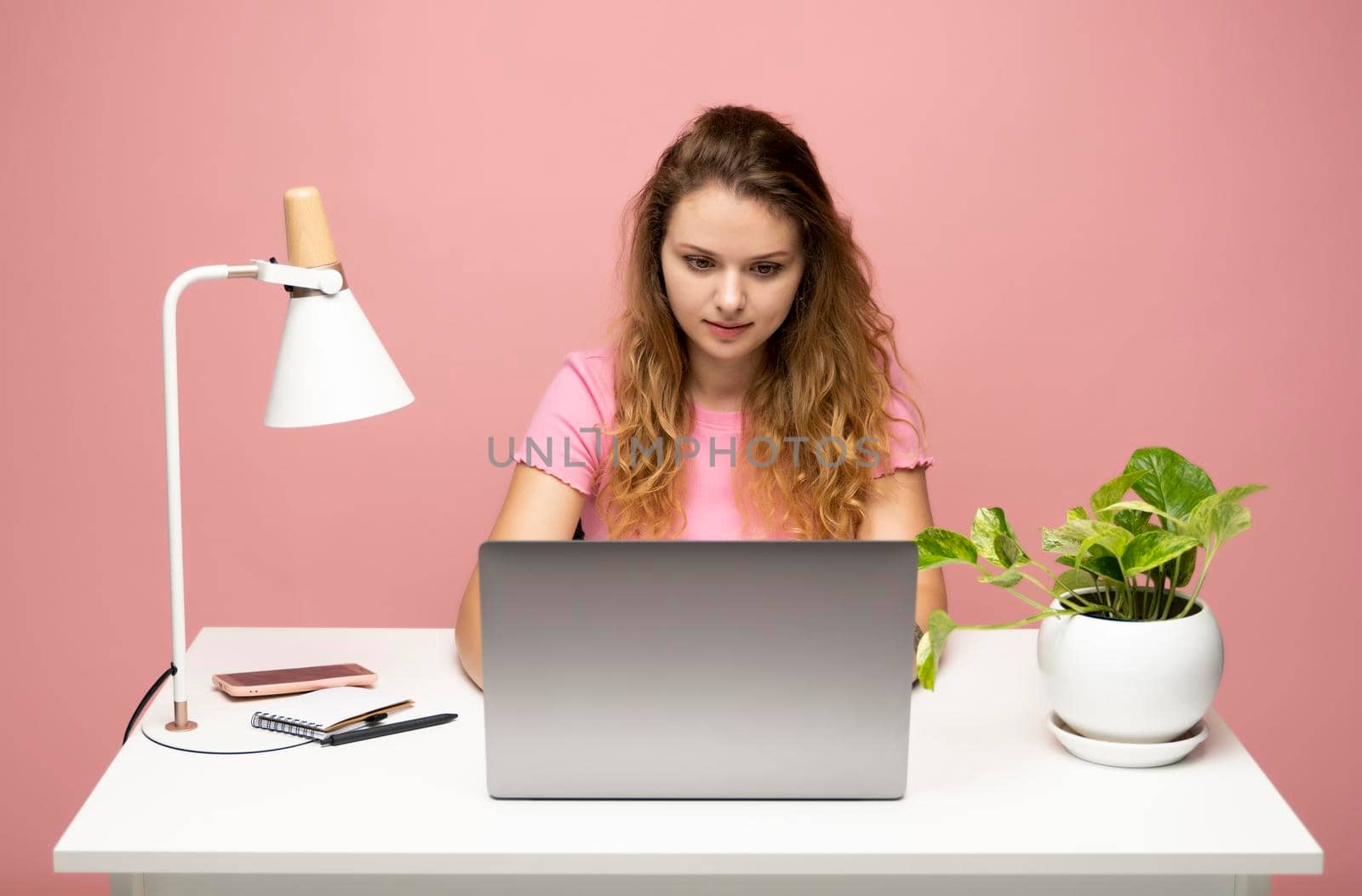 Young freelancer curly woman in a pink t-shirt working with a laptop computer over pink background. Working on a project. Freelance worker. by vovsht