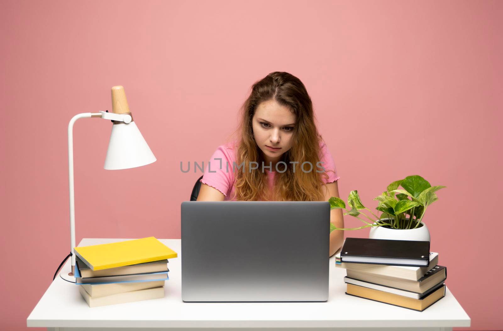 Young curly woman in a blue t-shirt studying from studio using laptop computer over a blue background. Serious beautiful young woman typing on laptop in a bright modern office