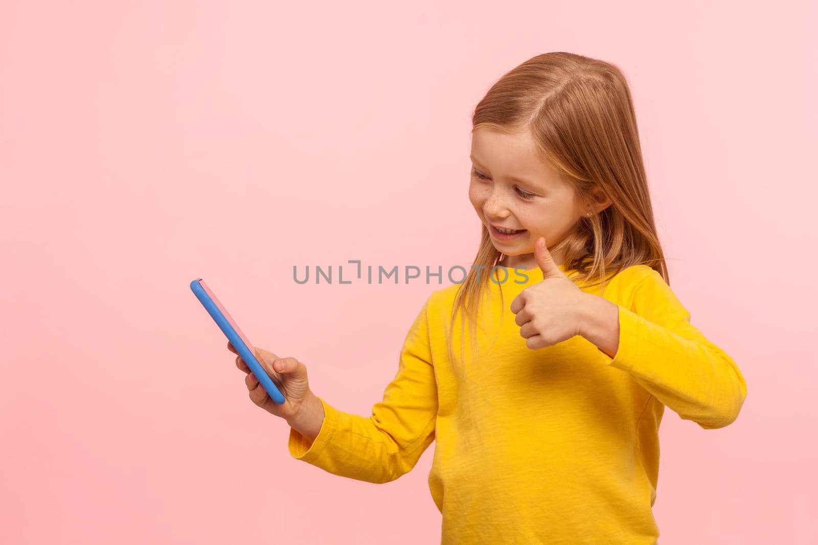 Portrait of excited little girl smiling and gesturing thumb up to mobile phone, showing like, approval sign while communicating with parents on video call. studio shot isolated on pink background