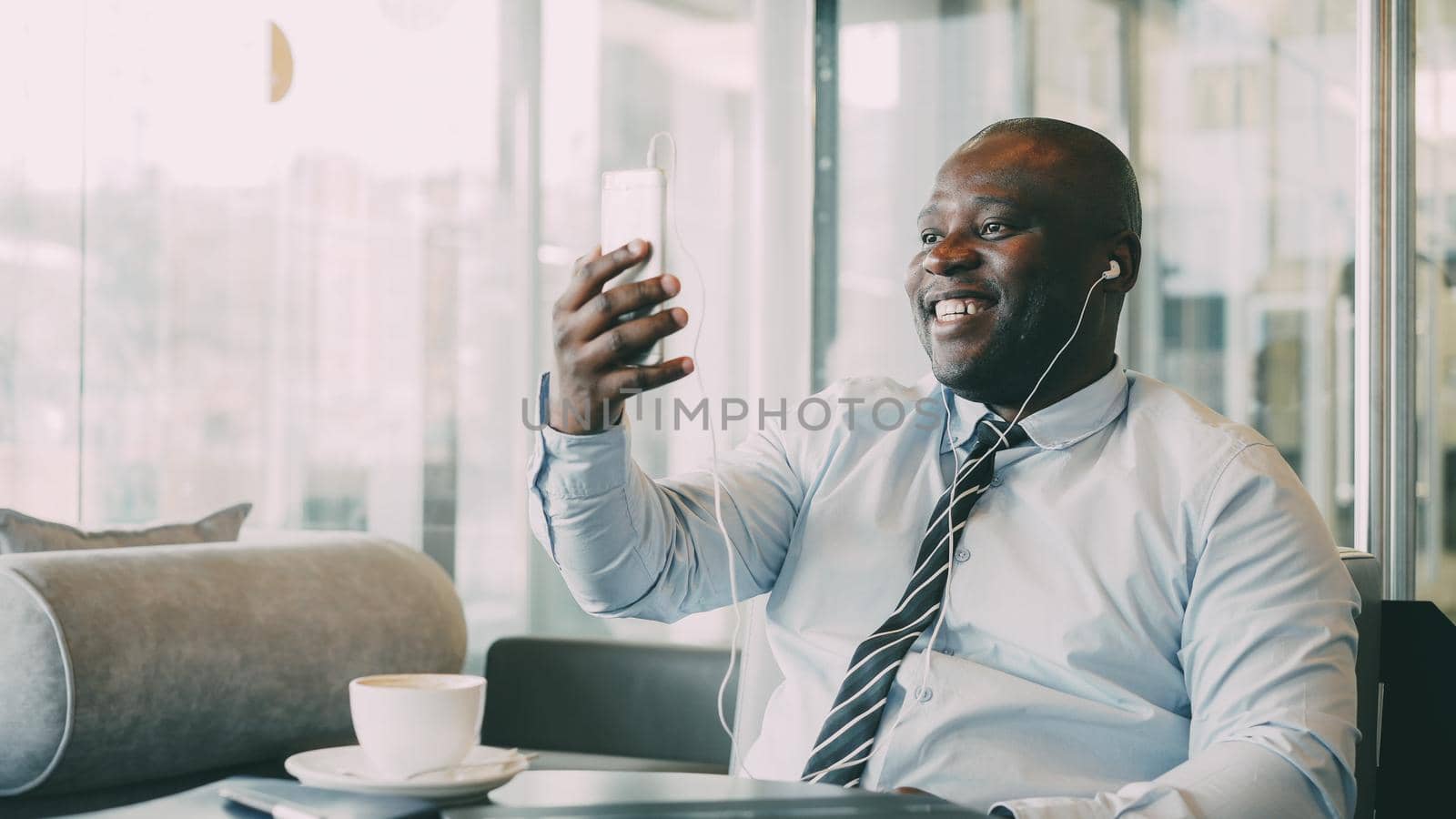 African American businessman in formal clothes laughing and talking to his family through smartphone video chat in airy cafe. He waves his hand cheerfully. by silverkblack