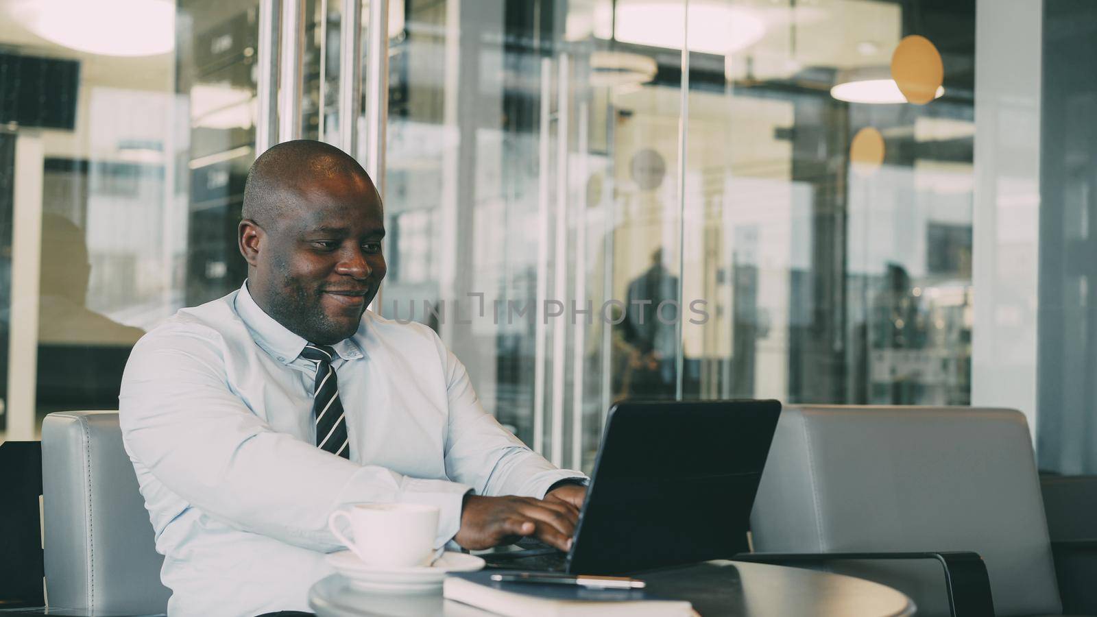 Cheery African American businessman smiling, printing and working on his laptop in glassy cafe during lunch break. by silverkblack