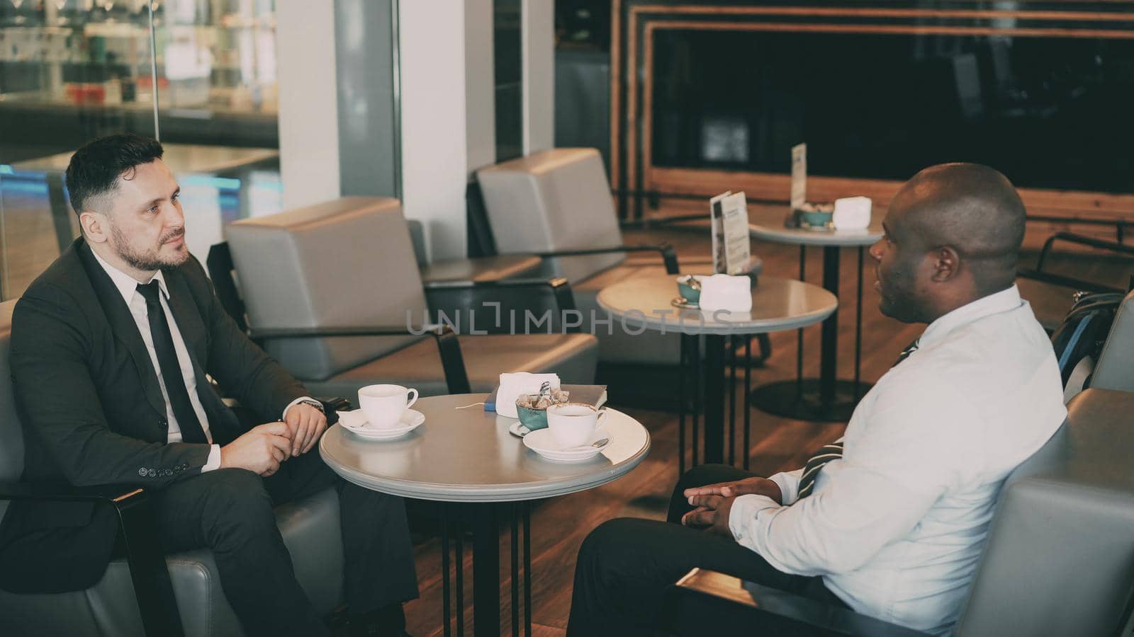 Two multiracial businessmen sitting, smiling and discussing their startup in airy cafe during lunch time. by silverkblack