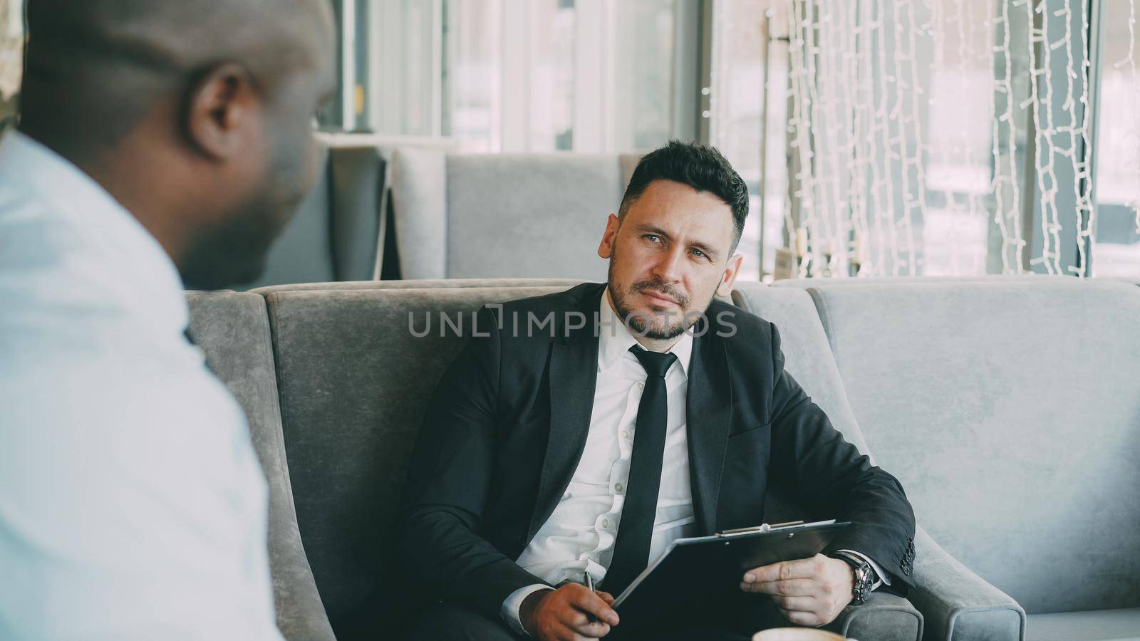 HR businessman having job interview with african american man and watching his resume application in modern cafe by silverkblack